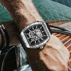 Luxury sport-inspired tonneau watch with skeleton dial on wrist, set against a yacht background